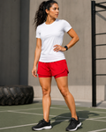 A woman in athletic wear posing on an outdoor court, wearing a white short-sleeve athletic t-shirt, bright red running shorts, and black running shoes