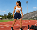 A fit woman stands on an outdoor running track wearing a white sports bra and navy athletic shorts, with stadium seating, trees, and a blue sky in the background. A black water bottle and bag sit on the track beside her.