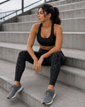A woman sits on concrete stadium stairs wearing a black sports bra and black leopard-print leggings with gray running shoes.